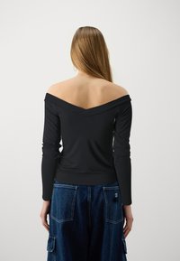 Black off-the-shoulder long-sleeve top with a V-shaped back neckline, crafted from a smooth fabric. Paired with blue denim cargo pants.