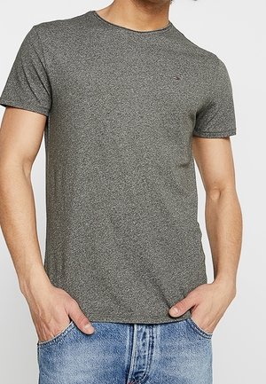 Mężczyzna noszący dopasowany, krótkim rękawem, szary melanżowy t-shirt z małym logo na piersi oraz jasno-niebieskie dżinsy, ręce w kieszeniach.