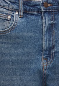 Vaqueros de mezclilla azul con diseño clásico de cinco bolsillos, botones de color cobre y costuras naranjas en contraste. Textura suave con un leve desgaste.