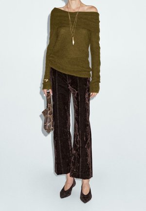 Femme portant un pull olive à épaules dénudées, un pantalon en velours marron foncé, des talons pointus, un long collier, un bracelet, et tenant un sac à main en cuir de serpent.