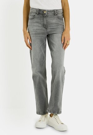 Personne portant un jean gris à jambes droites, des baskets blanches à plateforme avec lacets, une chemise blanche et une bague en or à la main droite.