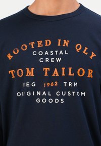 Tengerészkék ing narancssárga és fehér felirattal: "GYÖKEREDZIK A QLY-BAN", "TENGERI CSAPAT", "TOM TAILOR", "REG 1962 TRM", "EREDTI VÁSÁRLÓÁRU".