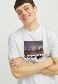 Weißes Baumwoll-T-Shirt mit einem grafischen Druck einer Palmenlandschaft und dem Text "Über Nacht." Das Shirt hat kurze Ärmel und einen Rundhalsausschnitt.