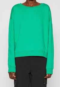 Sudadera verde, corta, con mangas largas y escote ancho. Fabricada en un material suave, con una textura lisa y puños acanalados.