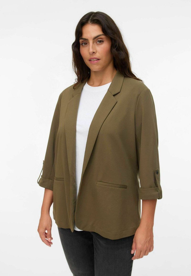 Olivgrüner Blazer mit lockerer Passform, hochgekrempelten Ärmeln und zwei vorderen Taschen, kombiniert mit einem weißen T-Shirt und dunklen Jeans.