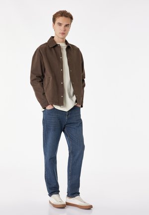 Joven de pie con las manos en los bolsillos, vistiendo chaqueta marrón, camisa blanca, vaqueros azules y zapatillas blancas sobre un fondo blanco.