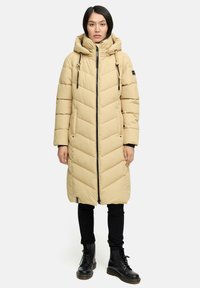 Beige pufferjakke med chevron-syning, hætte og snore. Frontlynlås og to sidelommer. Båret over sorte bukser og støvler.