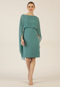 Tefarbene Kleid mit fließendem, durchsichtigem Überwurf. Figurbetonte Silhouette, knielang und weiche Textur. Paar silberne Sandalen mit Knöchelriemen.