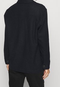 Musta villapaita-takki, jossa on kaulus, pitkät hihat, nappimansetit ja suora helma. Sileä pinta minimalistisella muotoilulla.