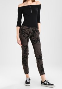 Svart av-axeln topp med knappar, kombinerad med mörka kamouflagejoggers med elastiska ärmslut, bärs med svarta sneakers.