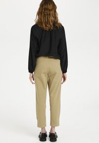 Svart långärmad topp med samlade manschetter, i kombination med beiga smala byxor och svarta skor, sedd från baksidan mot en vit bakgrund.