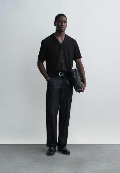 Άνδρας που στέκεται φορώντας μαύρο κοντομάνικο πουκάμισο με κουμπιά, μαύρο υφασμάτινο παντελόνι, μαύρα παπούτσια και κρατώντας μια μαύρη τσάντα μπροστά σε απλό τοίχο.