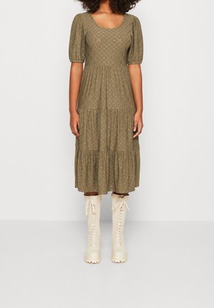 Femme portant une robe midi en dentelle ajourée vert olive avec manches bouffantes et bottes montantes à lacets crème, debout devant un fond uni.