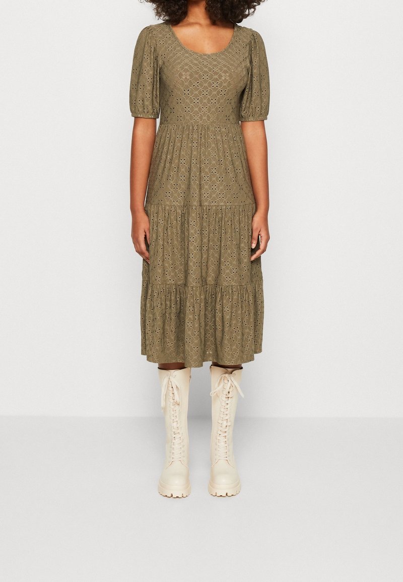 Femme portant une robe midi en dentelle ajourée vert olive avec manches bouffantes et bottes montantes à lacets crème, debout devant un fond uni.