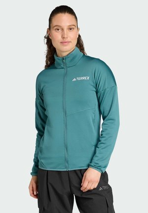 Femme aux cheveux tirés en arrière portant une veste zippée Terrex bleu sarcelle et un pantalon noir, debout devant un fond clair uni.