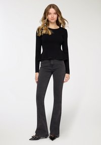 Top nero a coste con maniche lunghe e orlo a peplo, abbinato a jeans svasati grigio scuro a vita alta e décolleté nere appuntite con borchie.