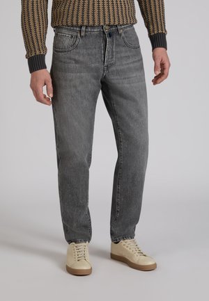 Uomo che indossa jeans grigi, sneakers beige e un maglione marrone e nero con motivo a fantasia, in piedi davanti a uno sfondo semplice.