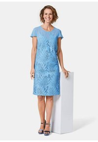 Himmelblaues Kleid mit kurzen Ärmeln, das filigrane ausgeschnittene Blumenmuster und eine glatte Textur aufweist, kombiniert mit passenden blauen Sandalen.