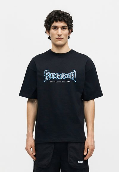 Νεαρός με κατσαρά μαλλιά που φοράει μαύρο T-shirt Barrow με μπλε λογότυπο και κείμενο «Greatest of All Time», στέκεται μπροστά σε λευκό απλό φόντο.
