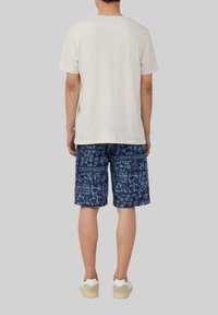 Lichtbeige katoenen T-shirt gecombineerd met blauwe korte broek met een bloemenpatroon. Model draagt witte sneakers met een lichtbeige zool.