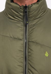 Olijfgroene gewatteerde jas met hoge kraag, zwarte rits en een klein groen logo op de borst. Glad textuur en lichte uitstraling.