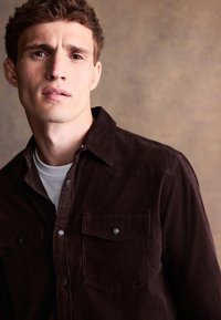 Brązowa koszula sztruksowa z kołnierzykiem, dwiema kieszeniami na piersi i zapięciami na zatrzaski, noszona na białym t-shircie. Teksturowana tkanina.