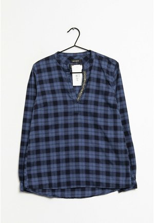 Blouse à manches longues à carreaux bleus et noirs avec un col en V et un détail décoratif le long de l'encolure, suspendue sur un cintre noir.