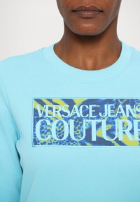 Világoskék kapucnis felső, amelyen egy élénk téglalap alakú grafika található, rajta a "VERSACE JANS COUTURE" felirattal fehér színben, valamint kék és sárga absztrakt mintákkal.