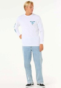 Langærmet hvid T-shirt med logo på brystet og turkis grafik. Matchet med lyseblå jeans og sorte sneakers. Afslappet pasform.