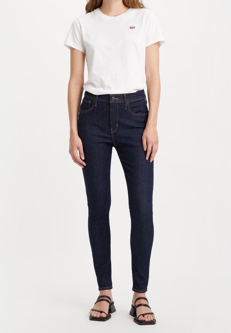 Jeans ajustados de color azul oscuro con costuras en contraste, cintura alta, bolsillos frontales, y una camiseta blanca con un pequeño logo rojo. Sandalias negras con tiras.