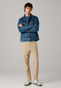 Jeune homme debout, les bras croisés, portant une veste en denim bleue, une chemise bleue, un pantalon beige et des baskets blanches, sur un fond uni.