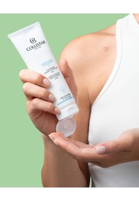 Crème nettoyante dans un tube souple blanc ; texte et logo bleus ; tenue dans une main avec une couleur d'ongle neutre ; arrière-plan vert.