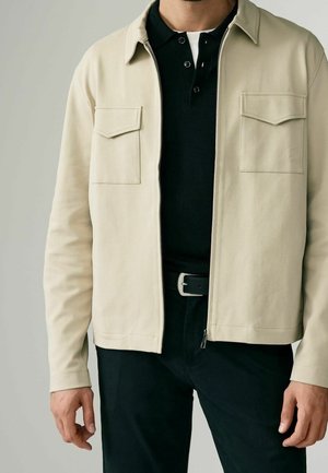 Homme portant une veste beige zippée avec des poches poitrine par-dessus une chemise noire à col et un pantalon noir avec ceinture.