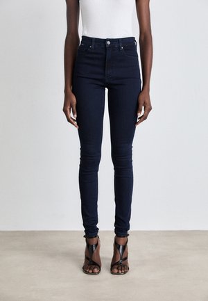 Jean slim bleu foncé porté par une personne debout portant un haut blanc et des sandales à talons hauts noires avec des brides.