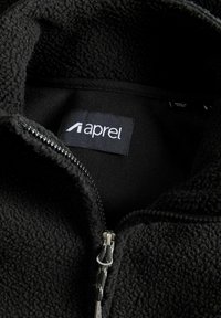 Veste en polaire noire à texture douce, dotée d'une fermeture éclair et d'une étiquette portant l'inscription "aprel". L'intérieur est doublé d'un tissu lisse.