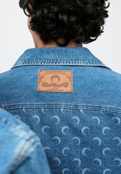 Világoskék színű farmerdzseki, texturált anyagból, barna bőrcímerrel a hátoldalon, amely félhold mintákat ábrázol.
