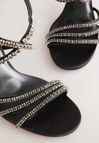 Črne sandale s peto z prepletenimi trakovi, okrašenimi z več srebrnimi kroglicami in kristali. Gladek črn podplat z mat zaključkom.