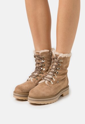 Jambes portant des bottes en suède beige à lacets avec doublure blanche pelucheuse, fermeture éclair sur le côté, et semelles en caoutchouc épaisses sur un fond blanc.