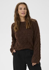 Pulover tricotat maro cu un model în diamante, având un decolteu rotund și mâneci ușor supradimensionate. Asortat cu pantaloni negri.