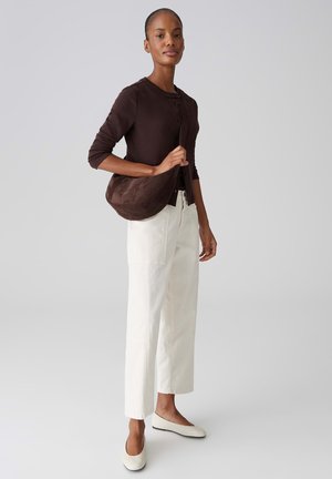 Frau trägt eine dunkelbraune Strickjacke, weiße Hosen mit weitem Bein, weiße Ballerinas und eine dunkelbraune Schultertasche auf ihrer rechten Schulter.