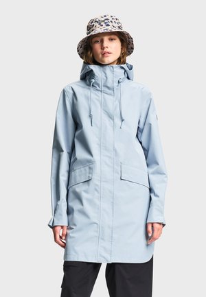 Ung person iført en lyseblå hættejakke og mønstret bøllehat, stående foran en ensfarvet lys baggrund.