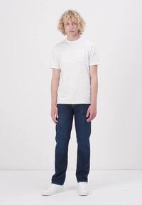 White cotton t-shirt with embossed "GUESS" logo, paired with dark blue denim jeans. Model stands against a plain white background.