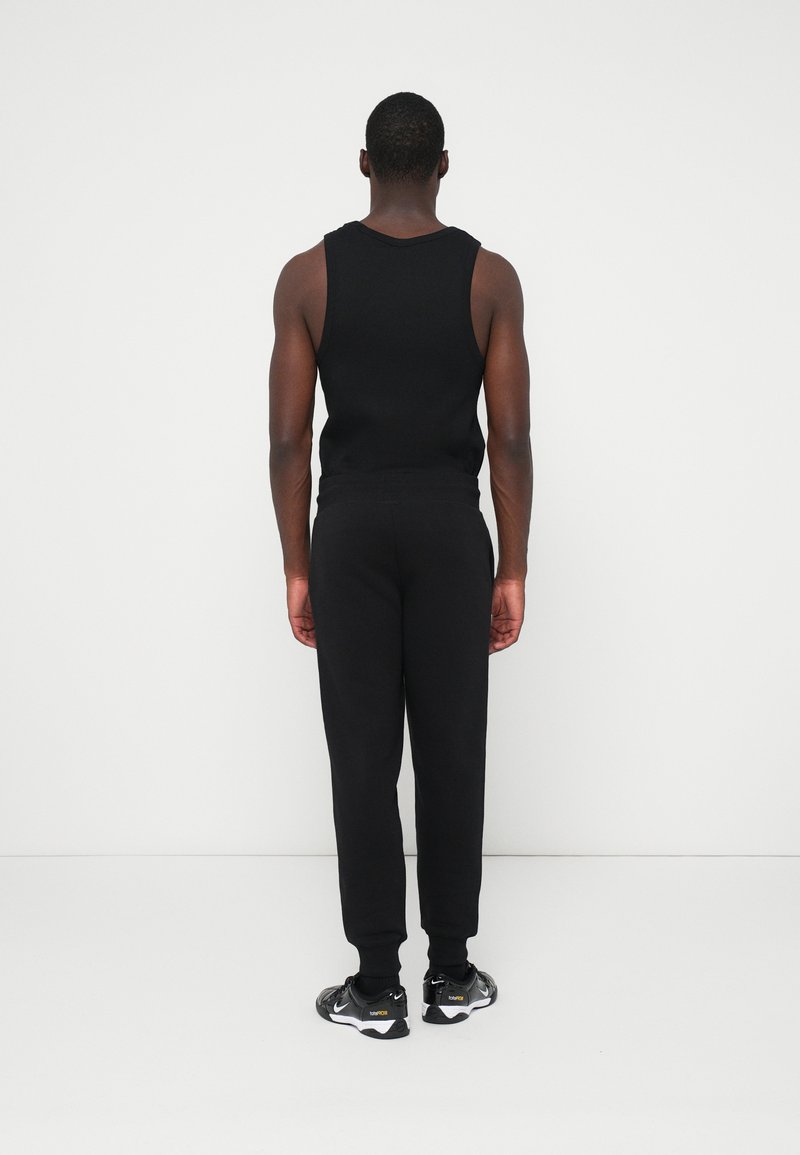 Black sleeveless tank top and matching joggers, both in a smooth, stretchy material. White-soled athletic shoes. Simple, minimalist design.