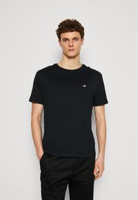 T-shirt en coton noir avec un col rond, des manches courtes et un petit logo brodé sur la poitrine. Texture lisse, coupe décontractée.