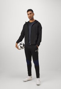 Zwarte sportjack met capuchon over een donker shirt, gecombineerd met zwarte broek met blauwe accenten. Witte voetbal en voetbalschoenen.