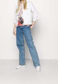Una persona se presenta con confianza, vistiendo una chaqueta blanca, una camiseta gráfica rosa y jeans azules, con zapatillas claras, sobre un fondo sencillo.