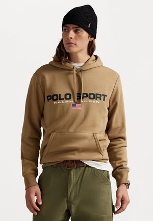 Tawny hoodie met zwarte "POLO SPORT" tekst, voorzien van een Amerikaanse vlag-embleem. Gecombineerd met olijfgroene broek en een gestructureerde beanie.