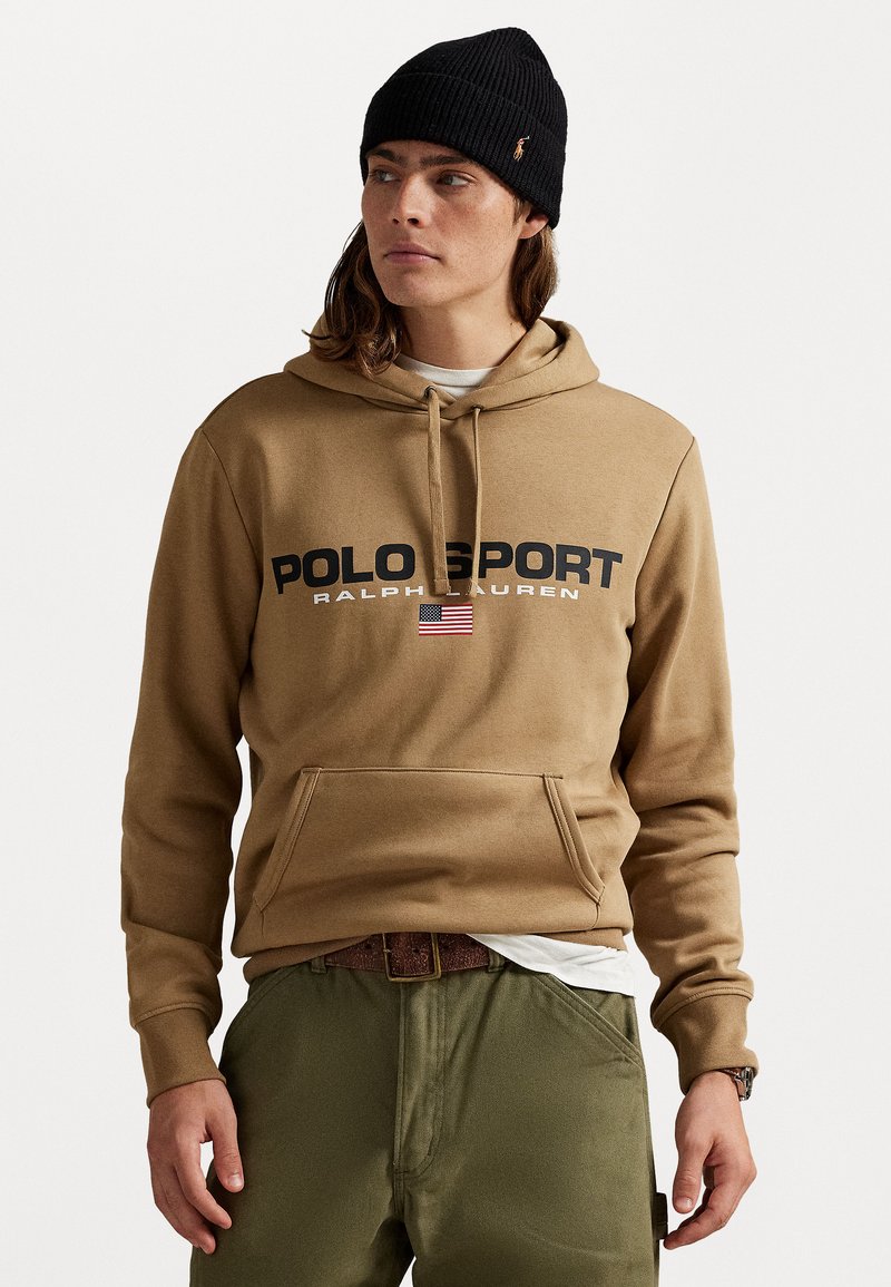 Hanorac cu glugă de culoare bej, inscripționat cu textul "POLO SPORT" negru, având un emblema a steagului american. Asortat cu pantaloni de culoare verde măslinie și o căciulă texturată.