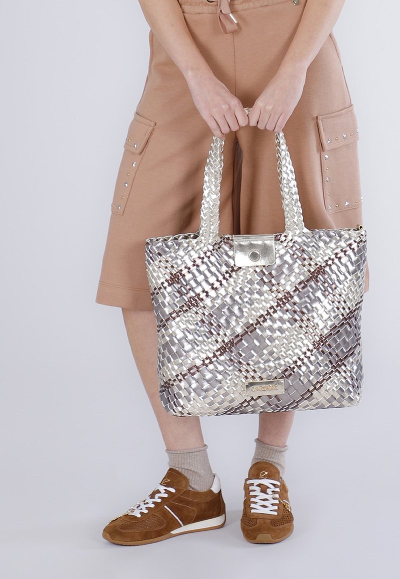 Persoon met beige korte broek en bruine sneakers houdt een grote zilverkleurige geweven draagtas met dubbele handvaten voor zich vast.