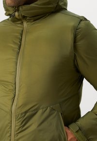 Veste à capuche vert olive avec une finition brillante, fermeture éclair sur le devant, poches latérales et accents au niveau des poignets en velcro. Présente une texture matelassée.